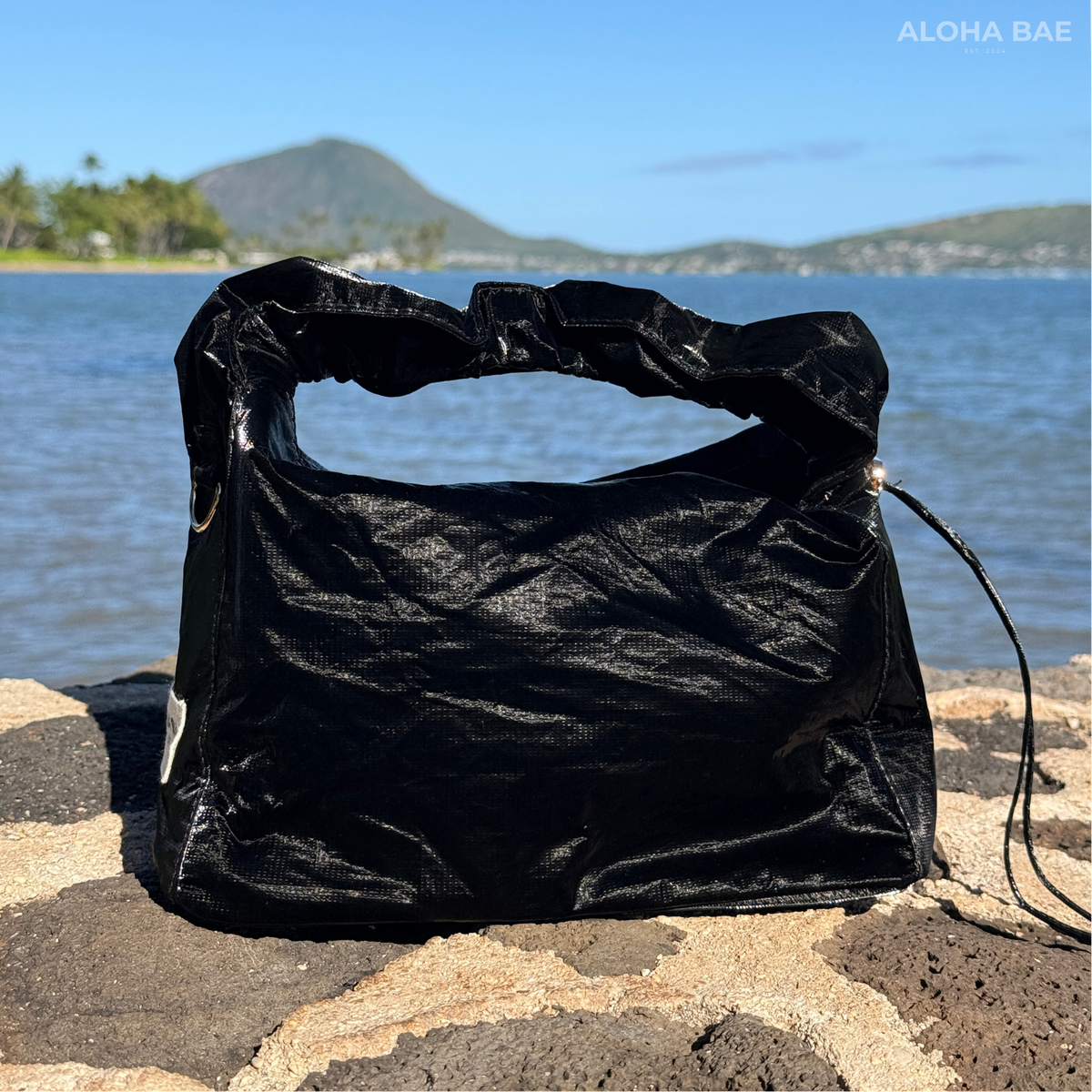 A black sheen hobo bag by Aloha Bae with a sleek, rounded design and shoulder strap, featuring stylish details and textured material.